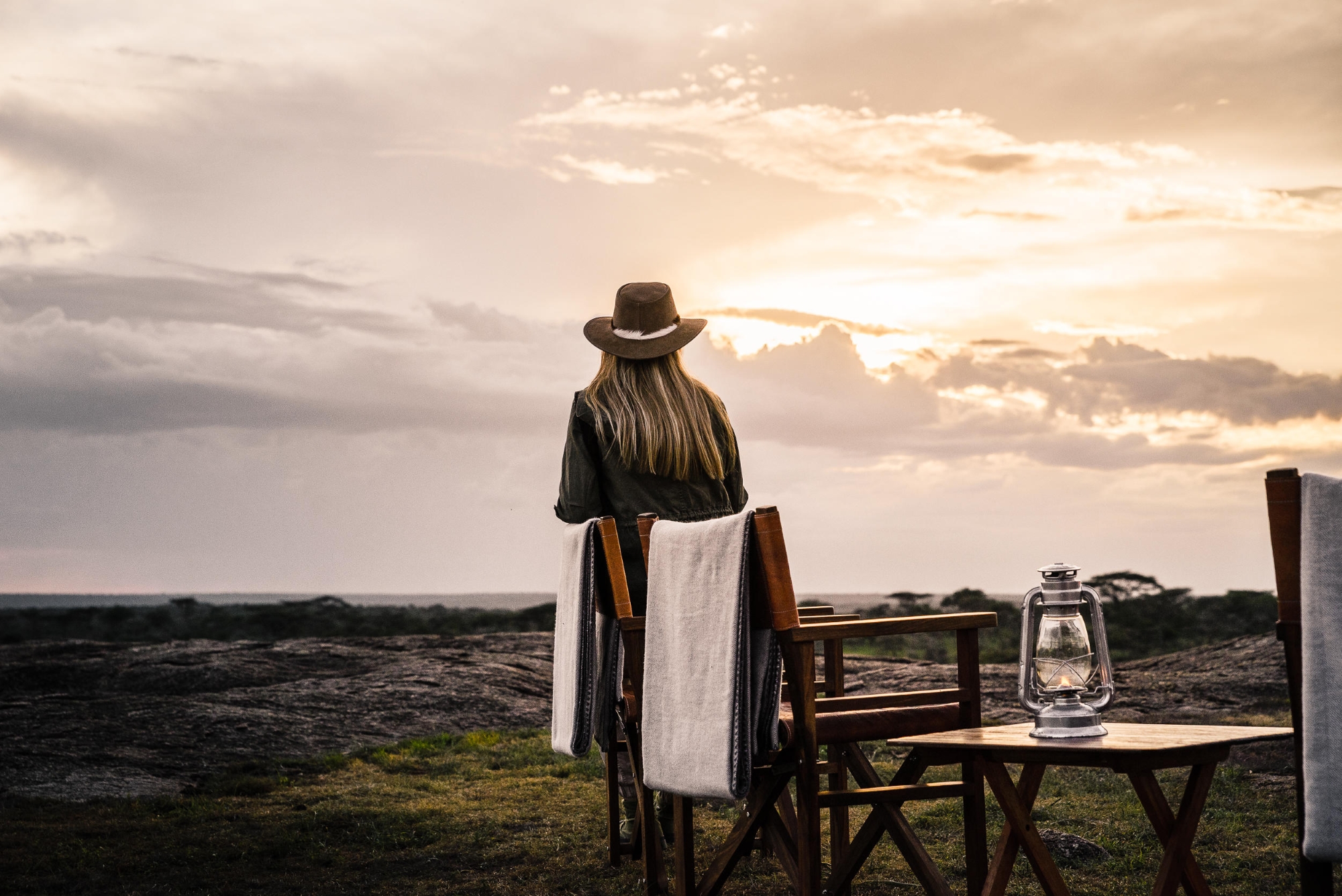 Kichakani-Serengeti-Camp-guest-sundown A woman in a safari hat sits facing the horizon at sunset, with empty wooden chairs and a lantern nearby on the grass