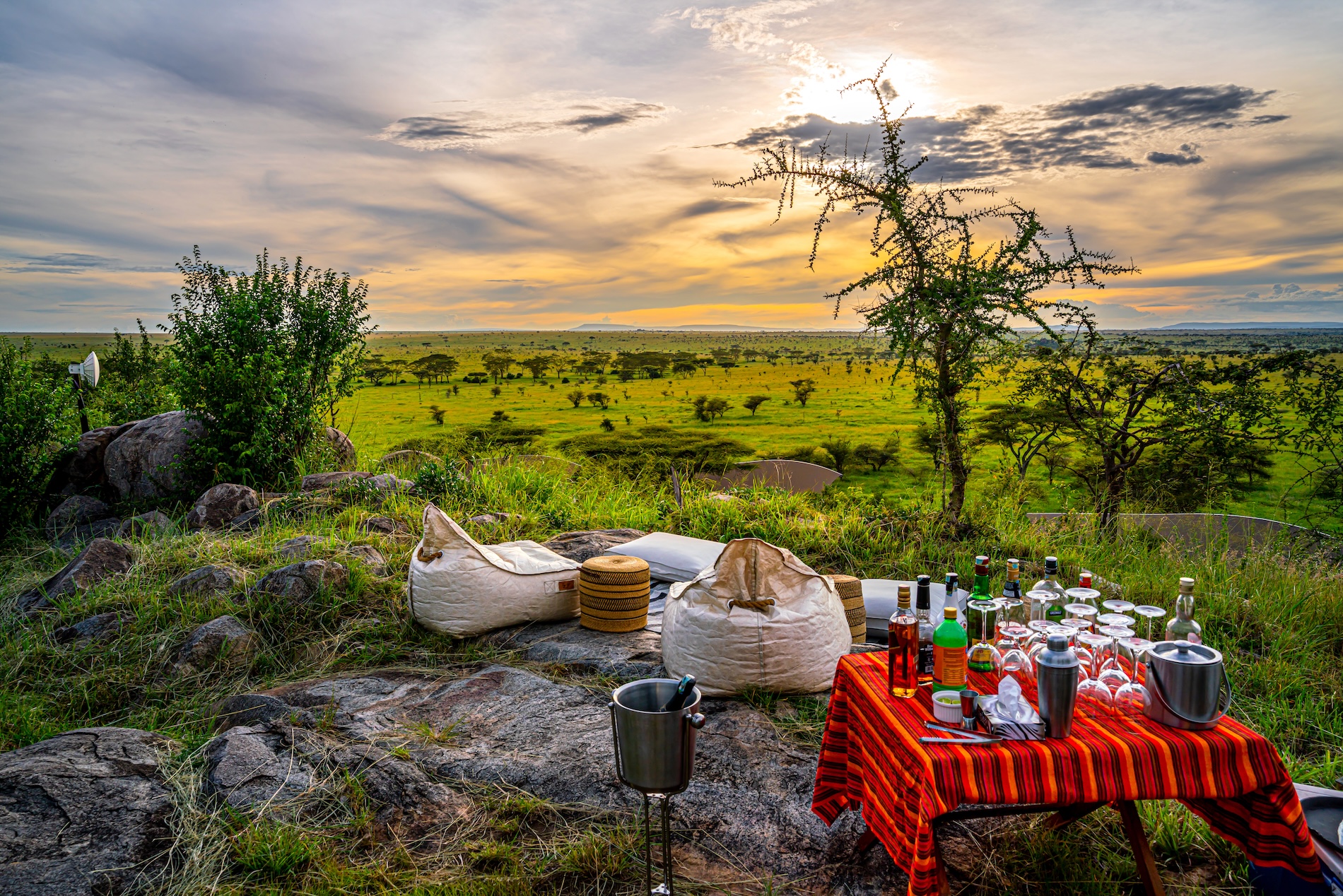 lemala-kuria-hills-lodge-sundowner-landscape-view-01 A scenic sundowner setup with beanbags and a table of drinks overlooks the vast green plains of the Serengeti under a golden sunset sky